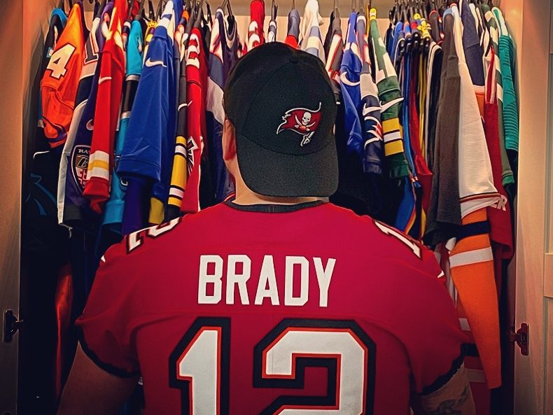 Eine Person steht in einem Tom Brady Trikot vor einem Kleiderschrank, der mit vielen anderen Sporttrikots gefüllt ist.