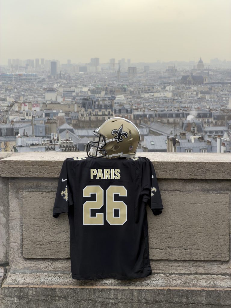 New Orleans Saints Jersey und Helm mit „Paris 2026“-Branding vor der Skyline der französischen Hauptstadt.